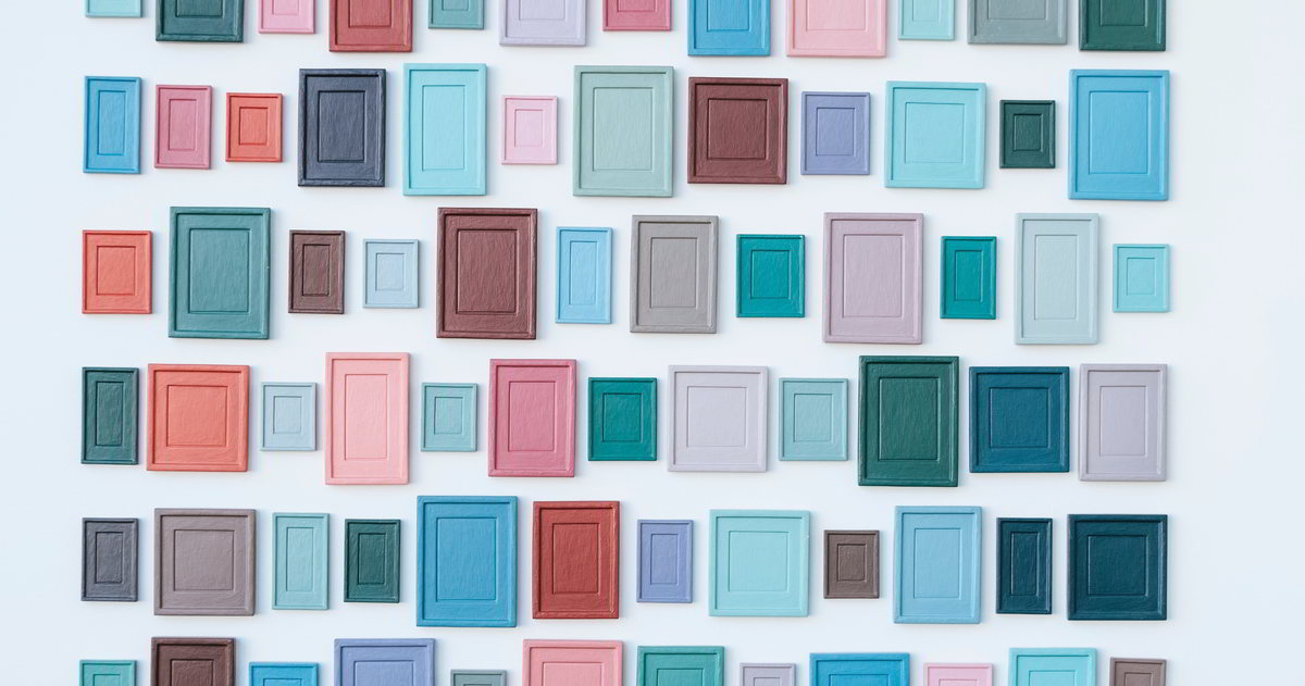In pink, t&uuml;rkis, helblblau, braun und gr&uuml;n angemalte Bilder verschiedener Gr&ouml;&szlig;en dicht an dicht an eine wei&szlig;e Wand geh&auml;ngt