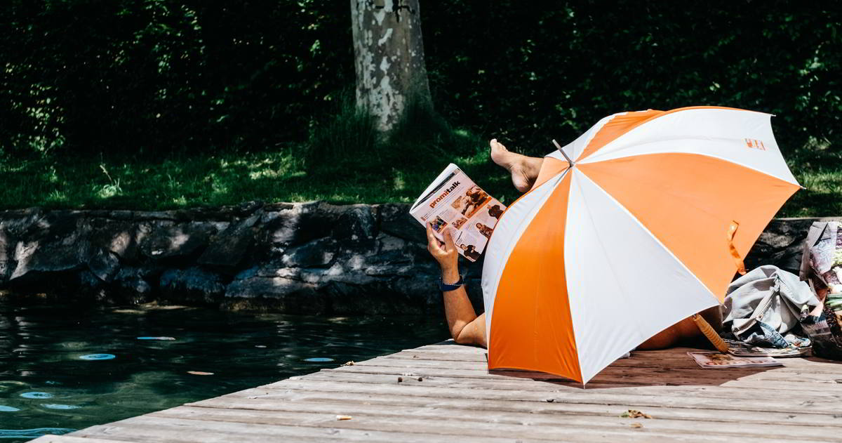 Entspannt lesen hinter einem orange-weiss gestreiften Schirm verborgen liegt ein Mann und auf einem Bootssteg und liest eine Zeitschrift